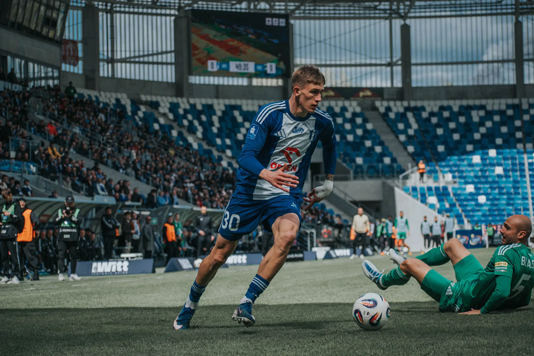 Piłkarz biegnie z piłką po murawie stadionu.